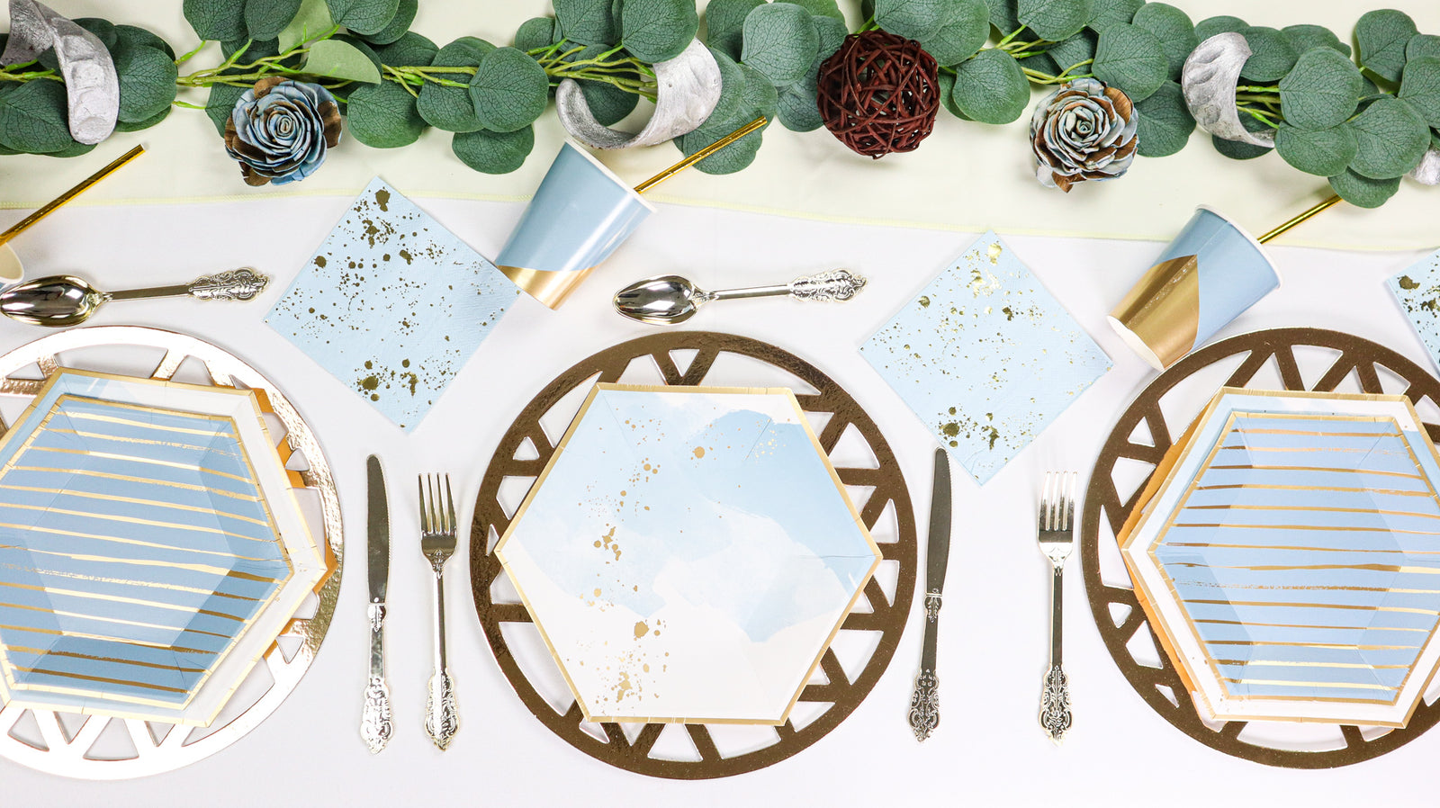 A set of party tables with golden blue hued plates and cutlery, arranged on a table with decorative flowers and a geometric pattern.