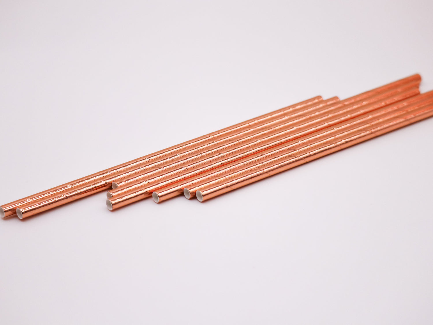 A set of metallic biodegradable paper straws in rose gold color, laid out on a white background.